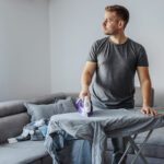 A man ironing his shirt in a single room occupancy (SRO) in front of a gray sofa and a white wall