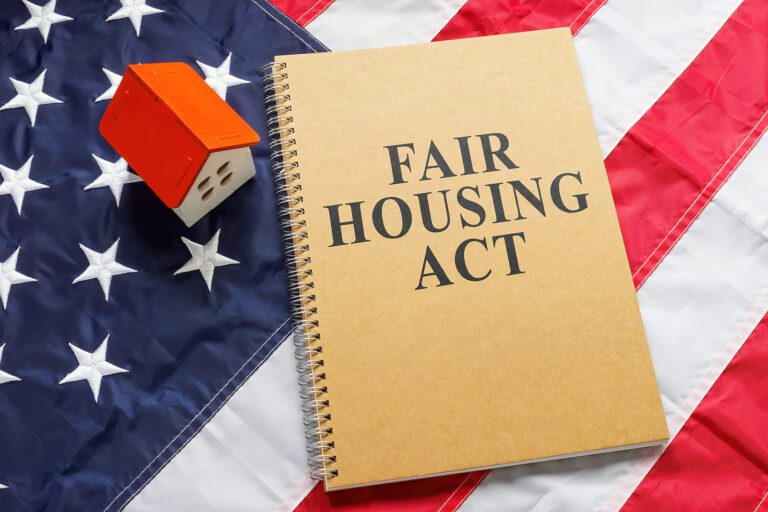 A notebook, with the text "FAIR HOUSING ACT" written on the cover, placed near tiny house on a US flag.